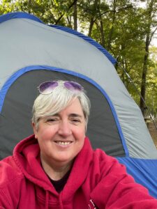 A woman with short gray hair and sunglasses on her head sits in front of a tent smiling