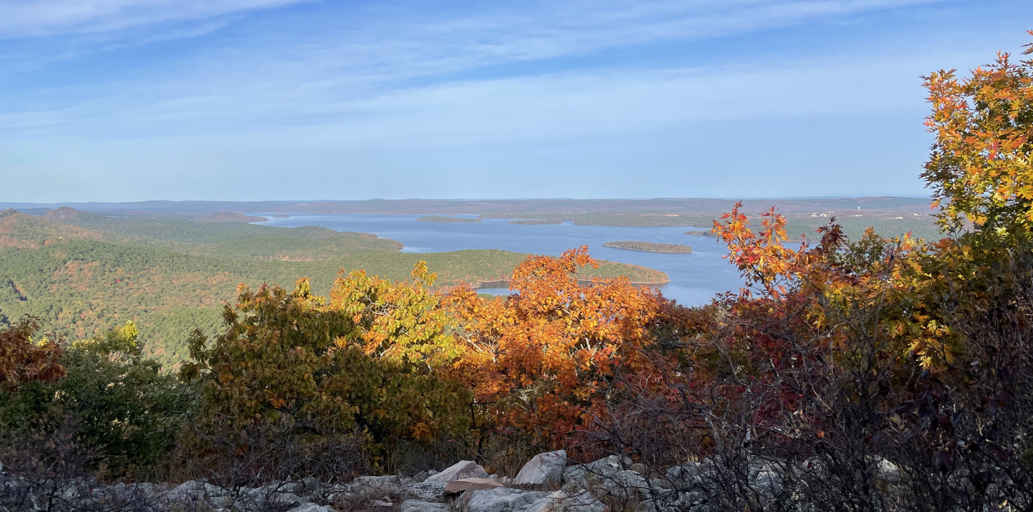 A view from the top of a mountain with rocks and orange and green trees nearby. In the distance is a large body of water surrounded by trees.