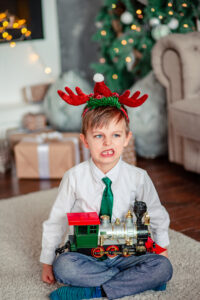 Child with reindeer hat on makes unhappy face as he holds a train in his lap