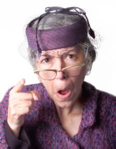 A woman with glasses and gray hair shakes her finger toward you with mouth open