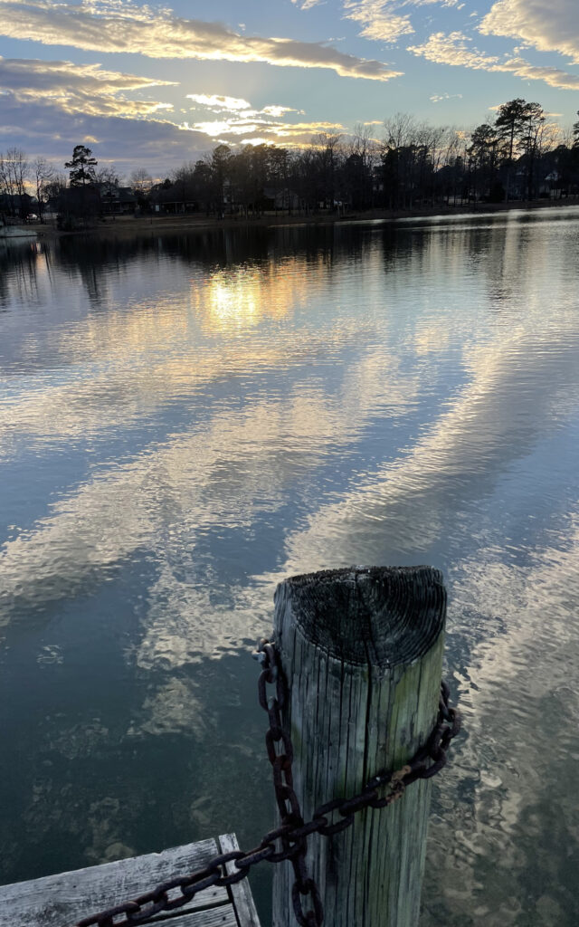 Standing on a fishing pier the post is nearby as we look out toward a tree-lined shore with light blue sky and bright white and yellow clouds. The colors of blue, orange, yellow and white from the sky are reflected in the water below.