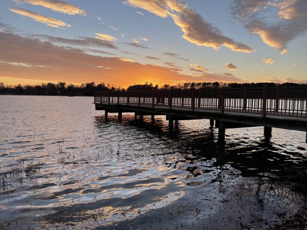 A pier stretches into a large body of water. On the horizon, there are clouds bright with orange and yellow floating in a blue sky. Hints of blue and orange are reflected on the rippling water.