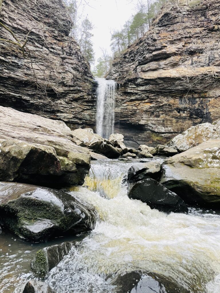 A water fall flows through a passage between two large cliffs. closer to us, water flows rapidly over several stones creating colors of white and yellow in the water.