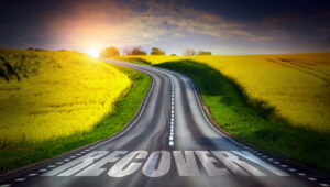 A road winds through a green meadow toward a sunset. The word recover is on the road in large letters.