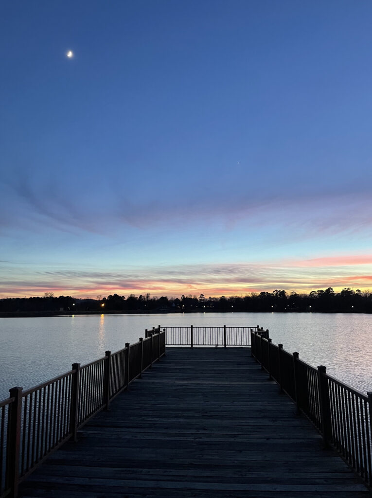 Looking out onto a fishing pier with a moon in the blue sky above and pink and light yellow clouds on the horizon beyond the lake