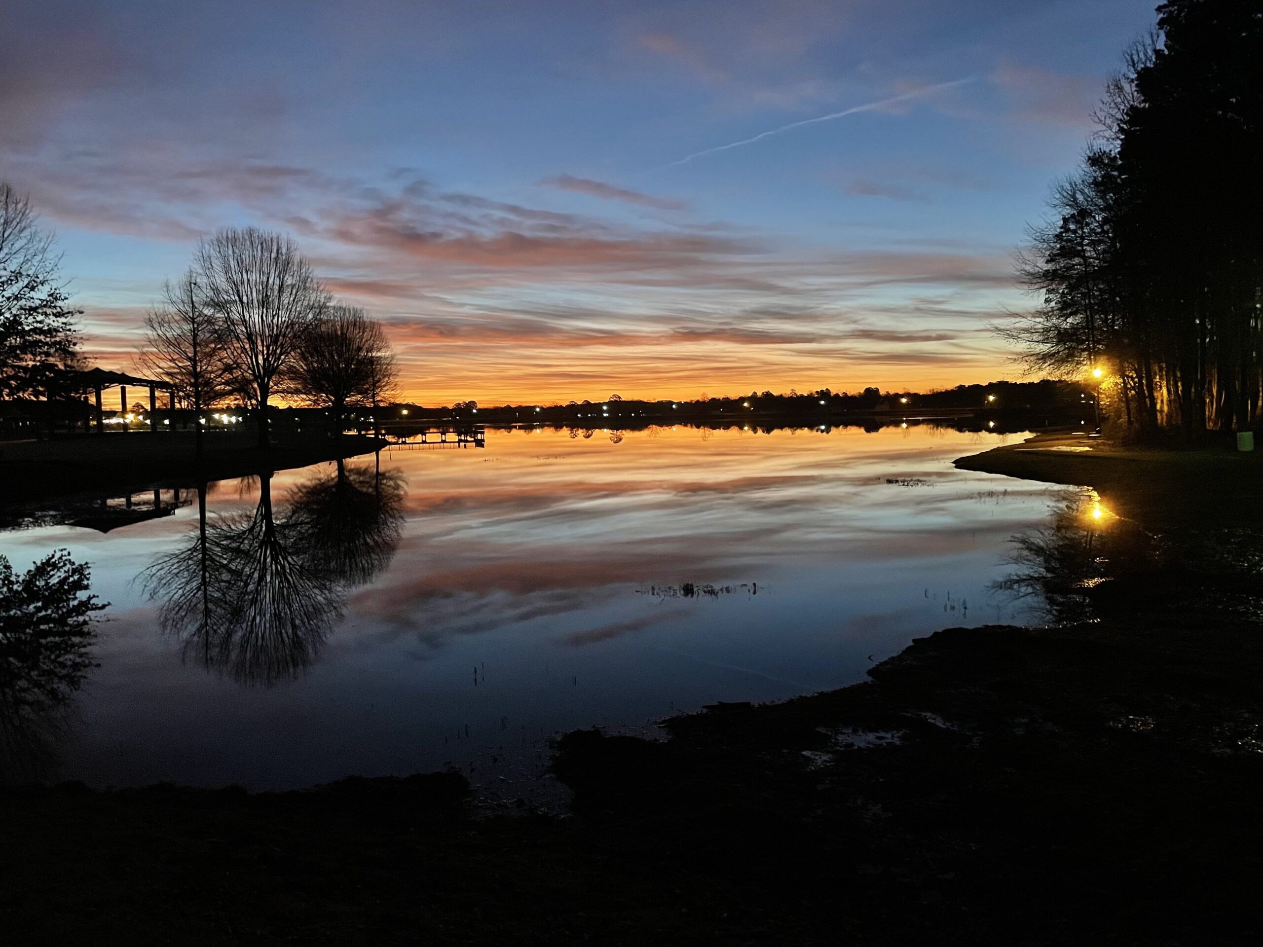 Early light breaks the horizon spreading orange color into a dark blue sky and reflecting in a body of water below. Silhouettes of trees line the shoreline. Street lamps can be seen across the lake.