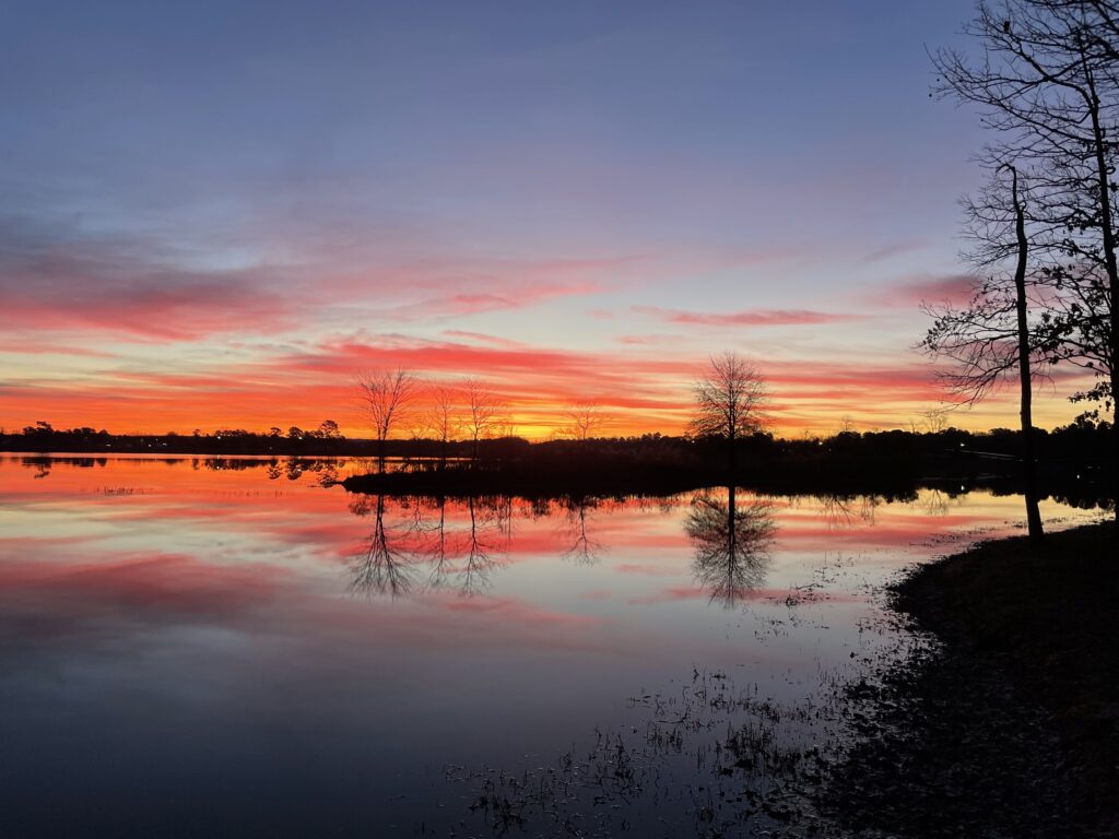 Bright red and orange clouds float on the horizon and fade into a blue sky above. Silhouettes of trees can be seen on the shoreline. The sky and trees are all reflected in the water below.