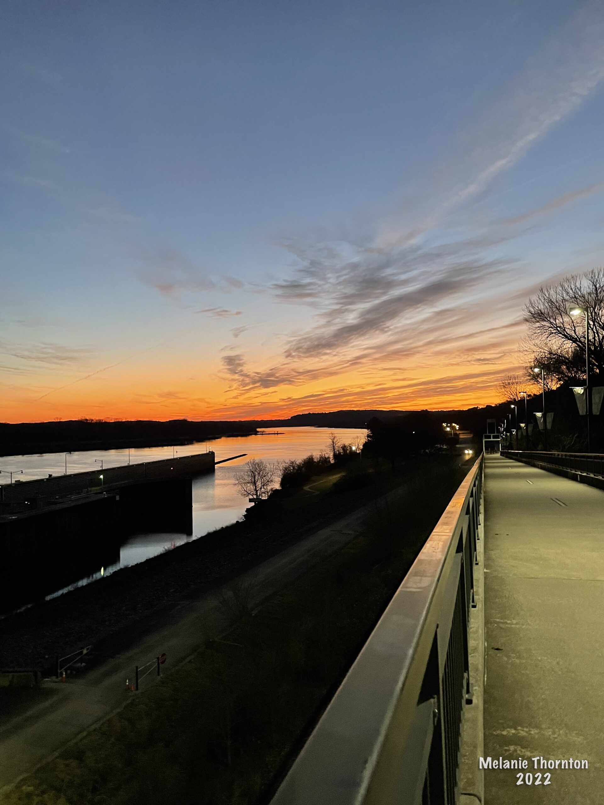 In foreground is a rail and a sidewalk. Looking over the rail is a body of water and a structure in the water. The sky is orange and yellow and blue.