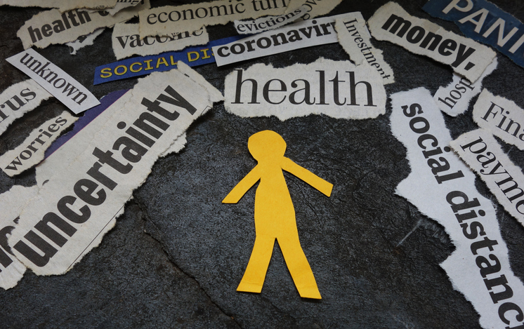 A paper cutout of a person is surrounded by words cut out of papers and magazines: social distancing, money, coronavirus, uncertainty, unknown, worries, health, economic, vaccine, evictions, investments