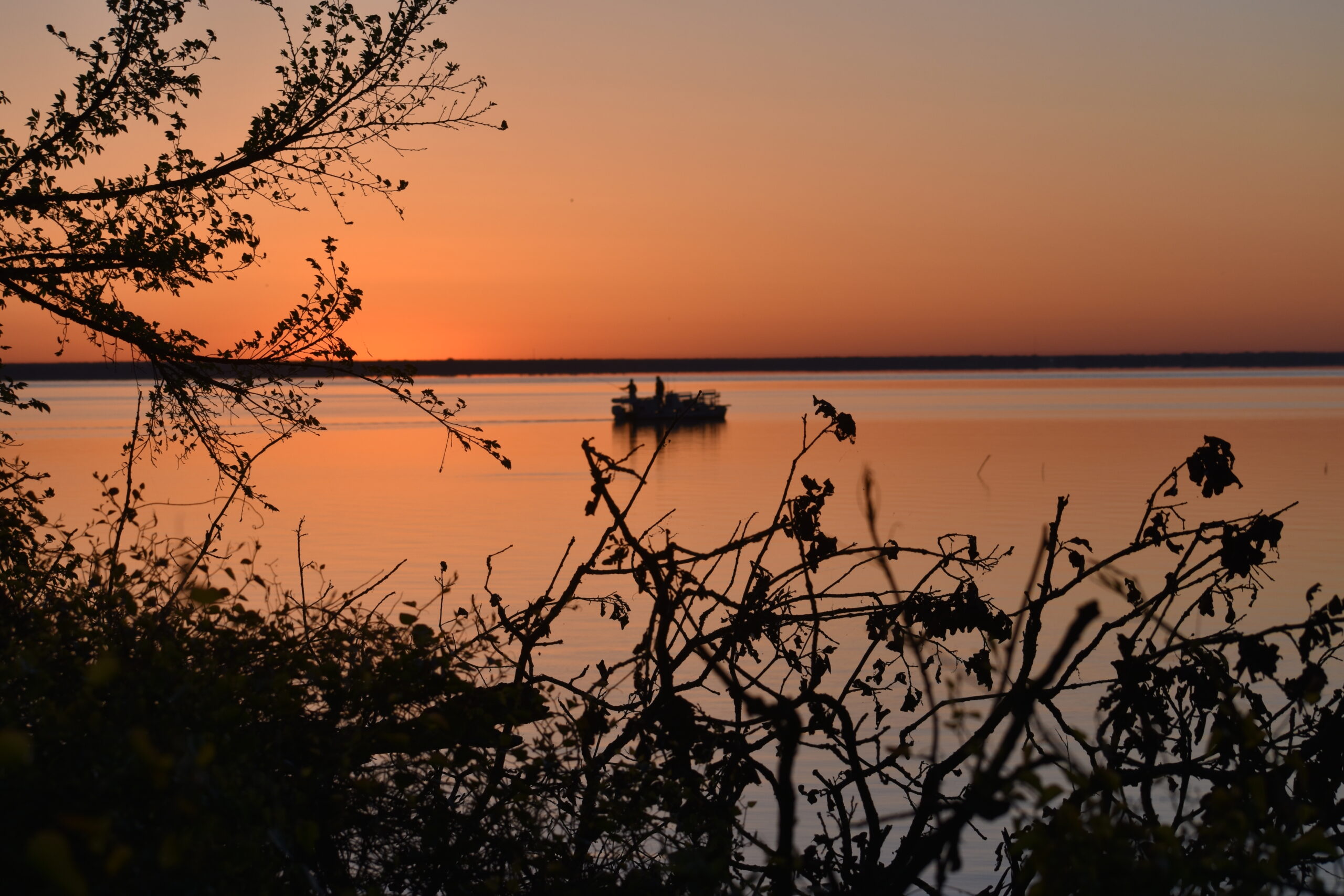Two men stand on a boat on the water of bright orange beneath a sky of the same color. Nearby on the shore are trees and bushes silhouetted by the bright orange water