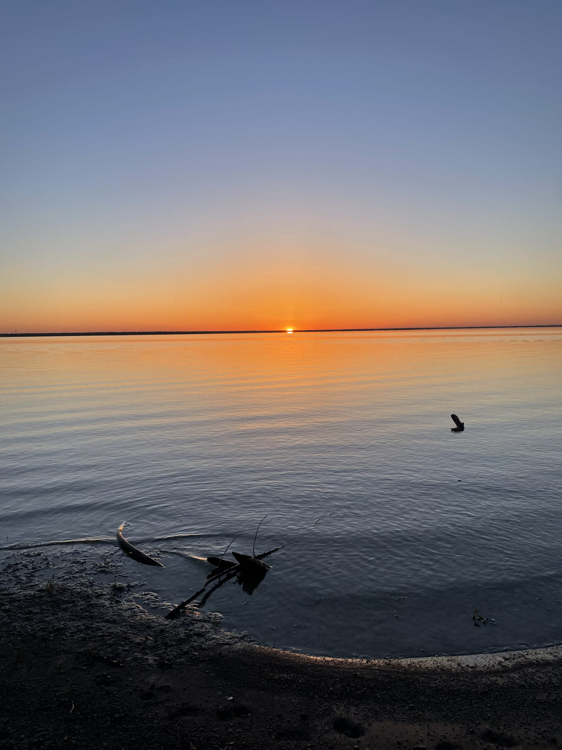 A tiny dot of a yellow sun sits on the horizon. The sky and water just above and below it are awash with bright orange. The sky fades from orange to yellow to blue above. The sandy shoreline is seen nearby.