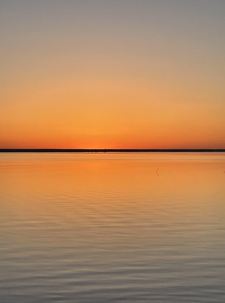 A thin horizon of black separates a bright orange sky above reflected in the bright orange water below.