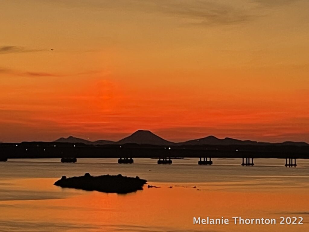 Several mountains in the distance are silhouetted by a bright orange sky. Closer to us a bridge extends across a large body of water which is orange with the color of the sky.