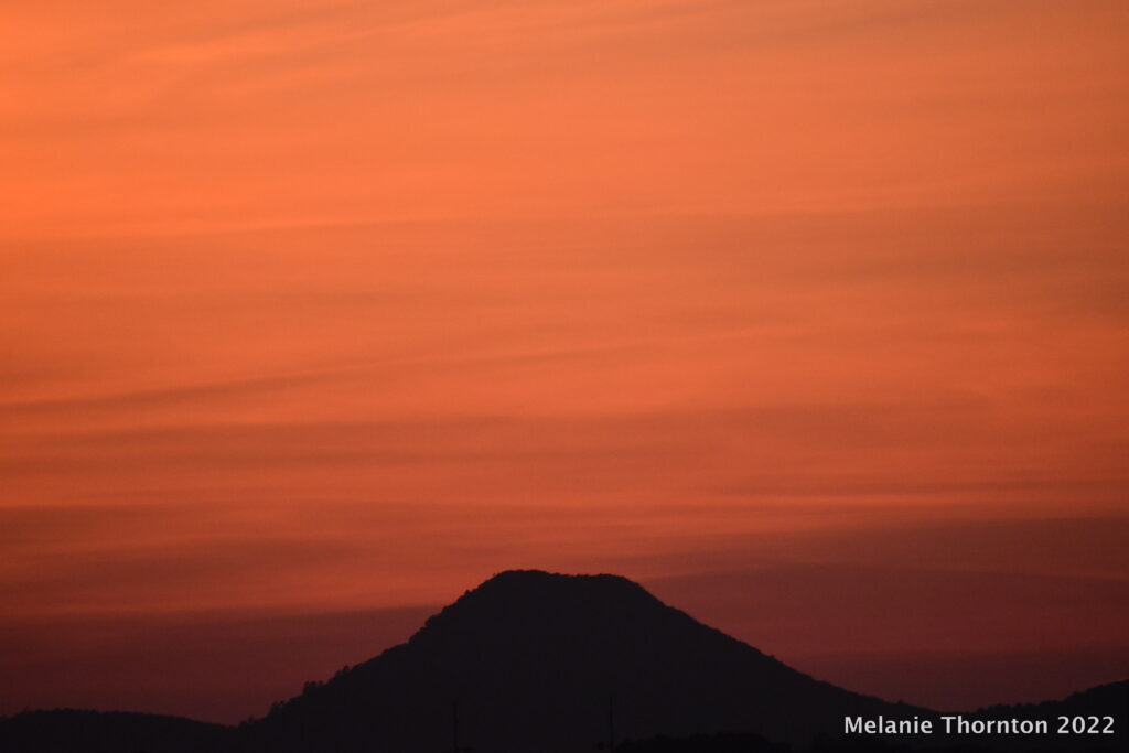 A mountain with a flat top is silhouetted by a bright orange sky that transitions from deep orange to brighter orange above.