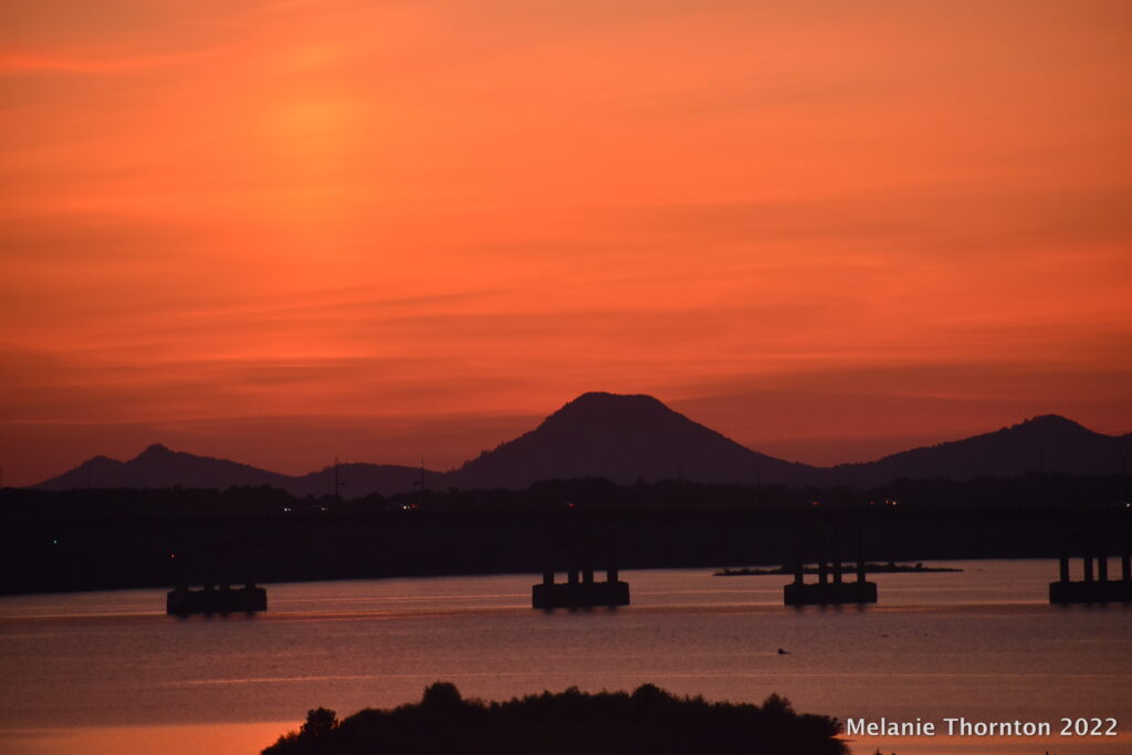 Looking across a large body of water, past a bridge, a row of mountains is silhouetted by a bright orange sky.