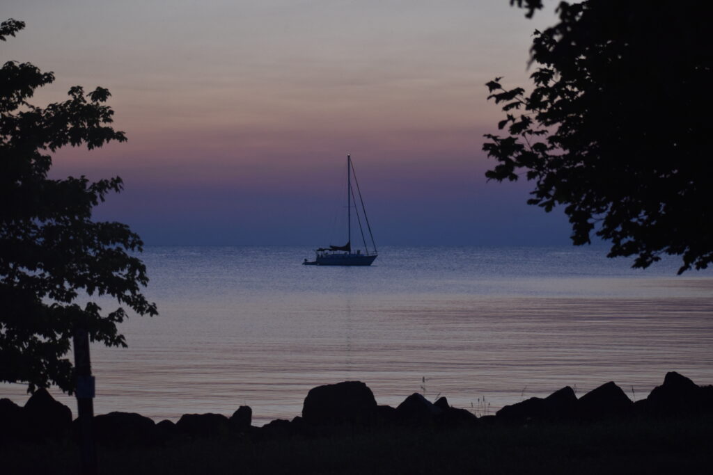 A black silhouette of rocks at the bottom and a tree on each side frame a sailboat. The purple and pink clouds behind it reflect in the water below.