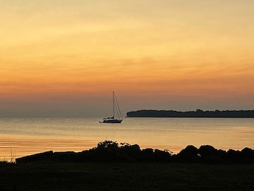 Looking out from a rocky shoreline, a sailboat rests in a harbor. Clouds of grey, orange and yellow are reflected in the water around it.