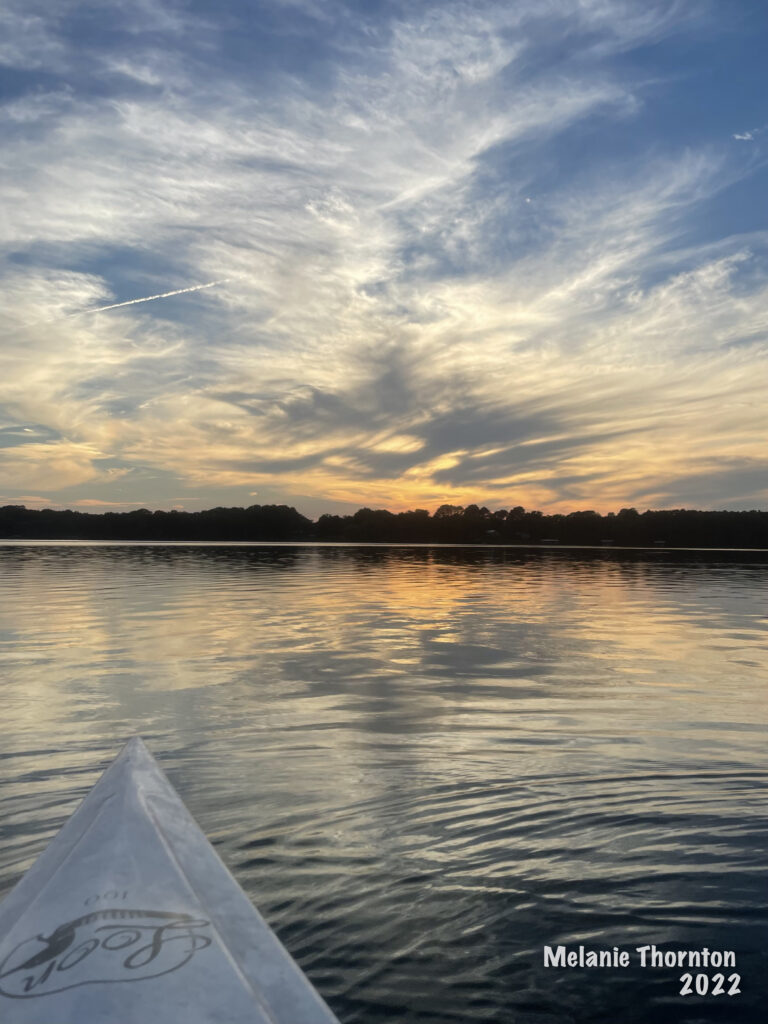 Looking over the nose of a kayak across a body of water filled with the flection of fluffy and wispy clouds. Toward the horizon the water is orange from the sunset above.
