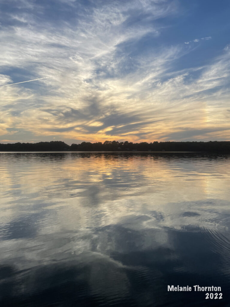 Looking across a body of water filled with the flection of fluffy and wispy clouds. Toward the horizon the water is orange from the sunset above.