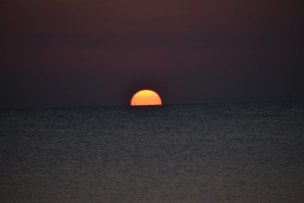 A dark photo with a bright round sun floating on the horizon on a large body of water. The sun appears to have soft stripes of light yellow at the top, transitioning to bright yellow, then orange, then red at the bottom. 