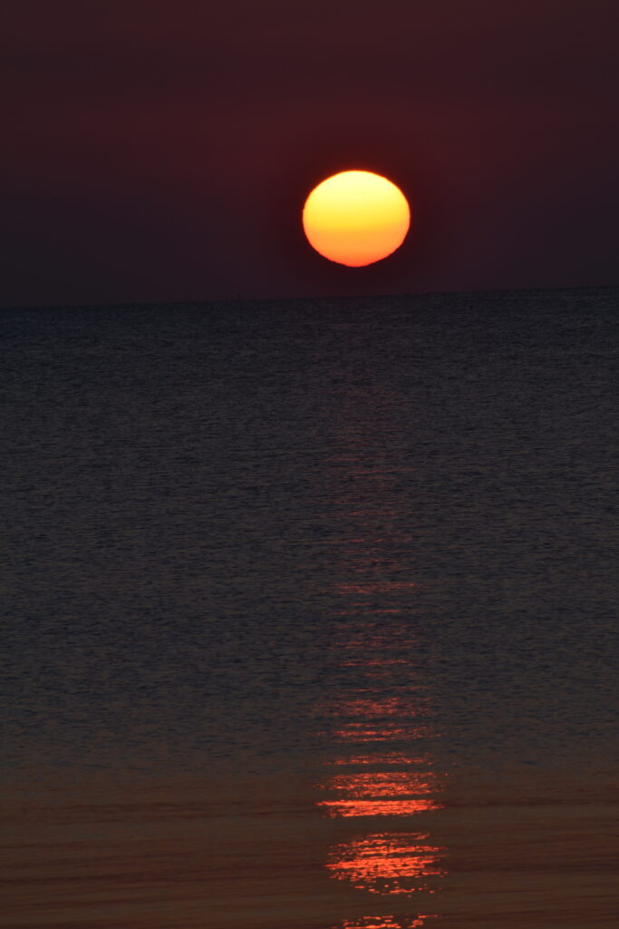 A dark photo with a bright round sun floating above a barely visible horizon on a large body of water. The sun appears to have soft stripes of light yellow at the top, transitioning to bright yellow, then orange, then red at the bottom. It casts a long reflection that gets brighter and brighter until it is bright red near us.