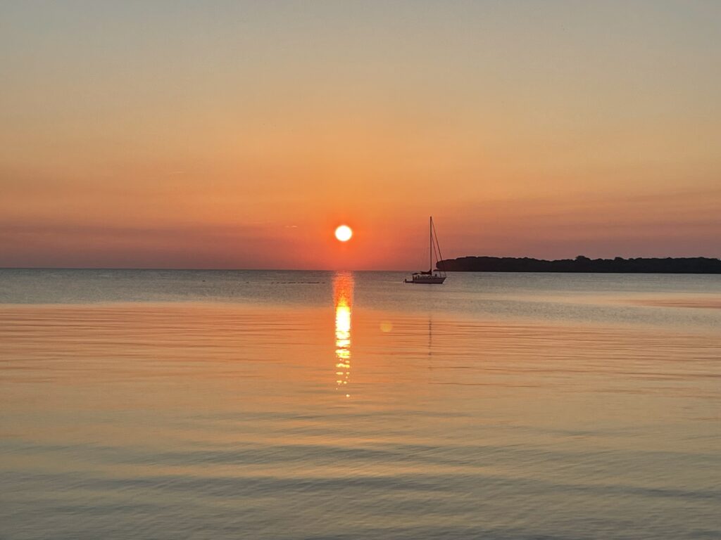 A bright orange sun sits just above the horizon and to the left of a sailboat on a large body of water. Bright orange color radiates through the sky and onto the water below.