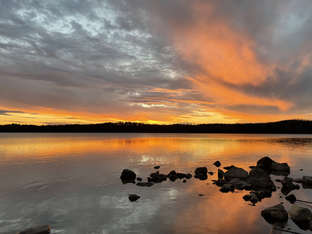 Looking over several large rocks that sit in the water, across a large body of water that is bright orange and yellow as it reflects the sky above. The clouds are grey above and bright orange on the lower side. The horizon is bright yellow and orange.