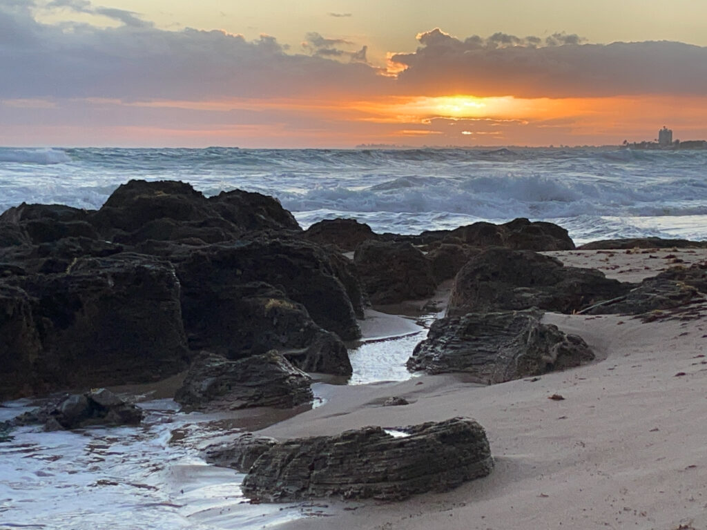 A bright orangish-yellow sun just above the horizon lights the sky and clouds with orange color contrasting against the blue-green water below. Closer, large brown rocks sit in the water and on the sandy shore.