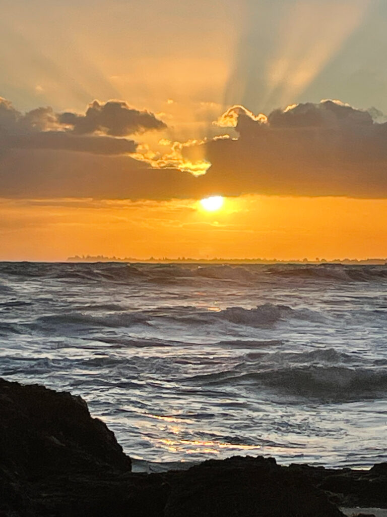 A bright yellow sun sits above the horizon tossing bright orange into the sky and causing the clouds to be lined in gold. Nearby, waves shimmer from the golden light near a row of dark rocks.
