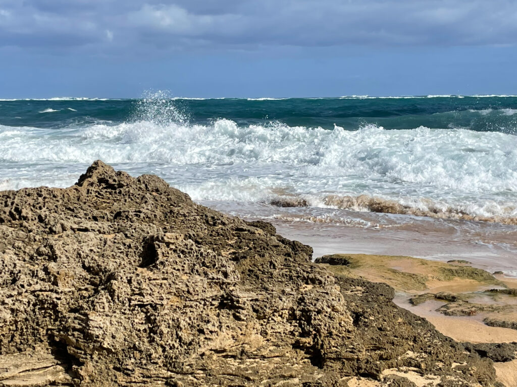 Looking over a large brown rock, waves crash against the shore. Beyond, the water is bright blue-green and the sky is grayish blue.
