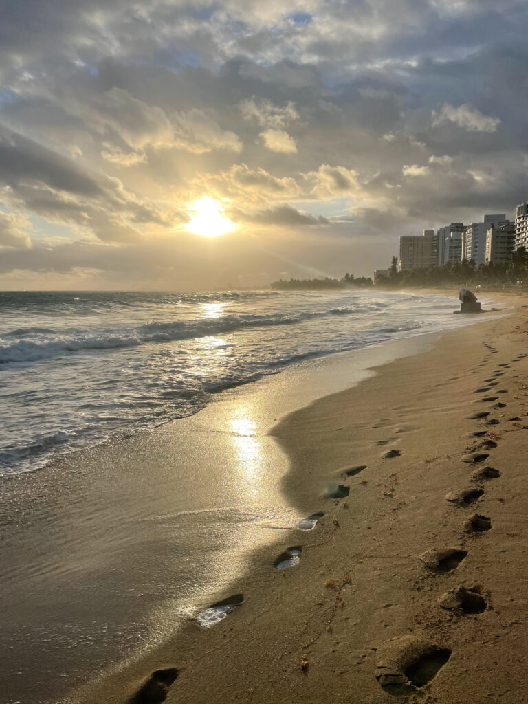 Two sets up footprints in the sandy beach stretch out toward a set of buildings on the shore. Waves move toward the short with a shimmering reflection of the sun which is just above the horizon peaking out from behind large fluffy gray clouds.